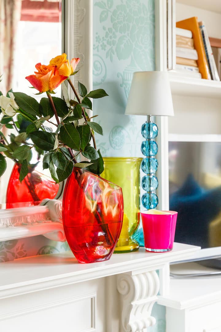 Photo détaillée d'une pièce montrant un mur avec du papier peint bleu clair, un miroir mural, et un manteau de cheminée blanc surmonté de chandelles, et un vase rouge avec des fleurs fraîches