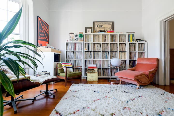 Un salon avec du parquet et de grandes fenêtres. Les points focaux principaux comprennent un tapis texturé et une grande collection de disques contre le mur du fond. Des affiches musicales encadrées sont accrochées aux murs, et il y a trois fauteuils rembourrés de différentes tailles et textures.