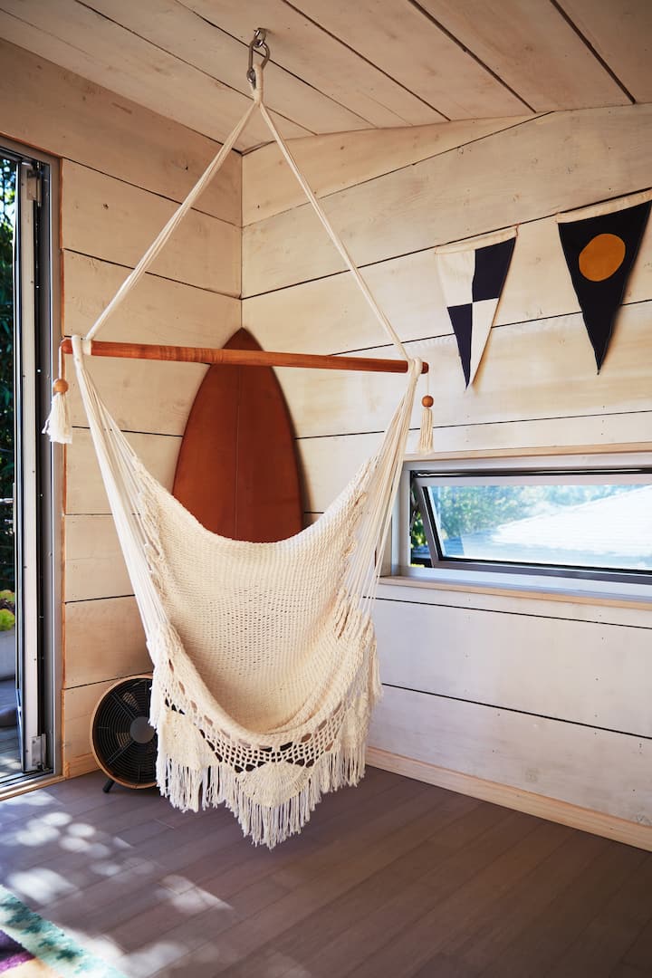 Une chaise-hamac tissée est suspendue au plafond près d'une petite fenêtre et d'une porte menant à un espace extérieur. Au-dessus de la fenêtre sur le mur de bois à droite pend une guirlande de drapeau triangulaire.