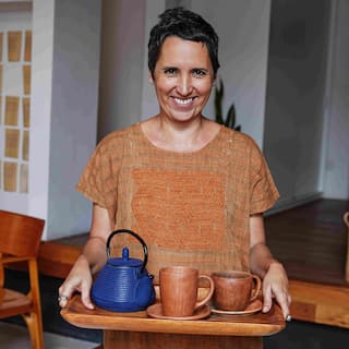 짧게 자른 갈색 머리의 여성이 찻주전자와 찻잔이 놓인 쟁반을 들고 미소를 지으며 서 있습니다.