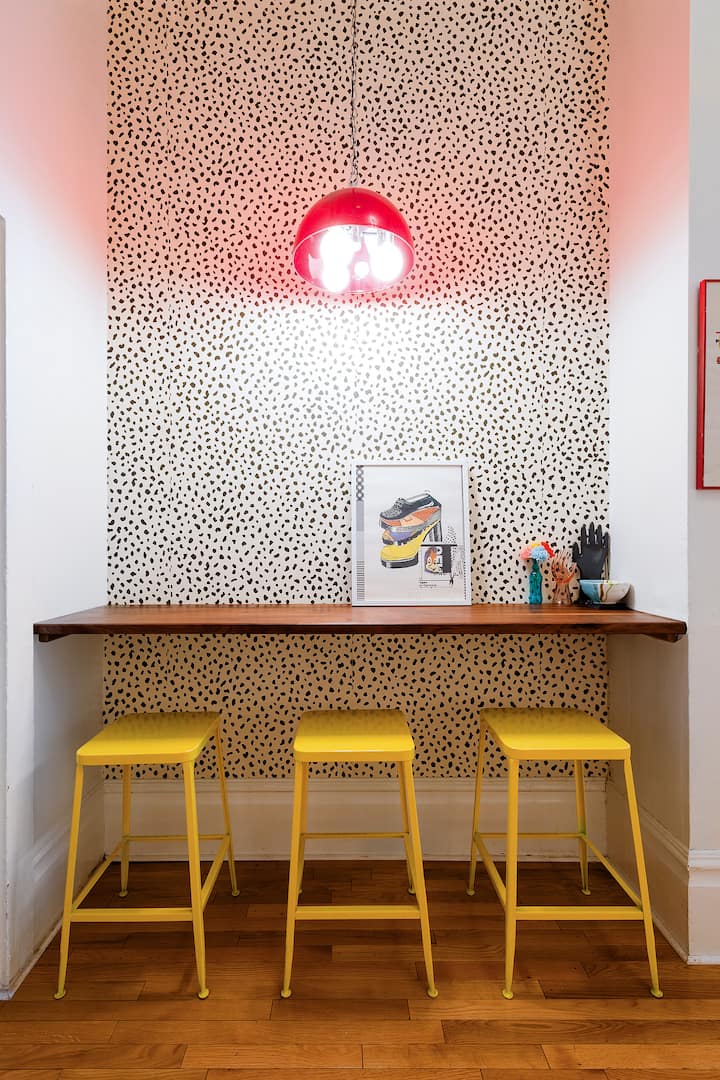 Un recoin éclairé par une petite lampe rouge. Le mur est orné d'un motif de papier peint noir et blanc. Une dalle de bois montée de part et d'autre du recoin sert de bureau, surmonté d'œuvres d'art encadrées et de petits objets et sculptures. Sous le bureau se trouvent trois tabourets en métal jaune.