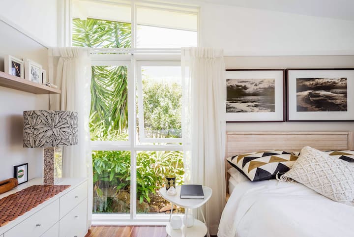 Coin lumineux d'une chambre avec des murs blancs, des œuvres d'art encadrées et une petite table de chevet. Les rideaux blancs recouvrent partiellement les fenêtres sol-plafond, dont une vitre est légèrement ouverte.