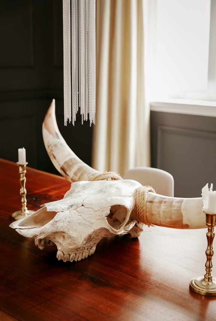 Une table en bois avec une pièce centrale décorative constituée par un crâne de vache partiellement enveloppé dans une corde de sisal et une paire de bougies en cire semi-fondue dans des chandeliers en laiton. Au-dessus du crâne se trouvent plusieurs brins de chaîne légère, ce serait éventuellement des éléments d'un plafonnier ou lustre. En arrière-plan, la lumière du soleil traverse la fenêtre habillée d'un rideau blanc.