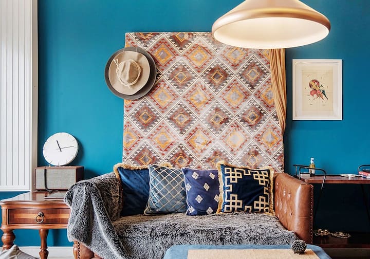 Un sofá de cuero marrón cubierto de cojines cuadrados y una suave manta descansa en una habitación azul. Cuadros y elementos rústicos visten las paredes.