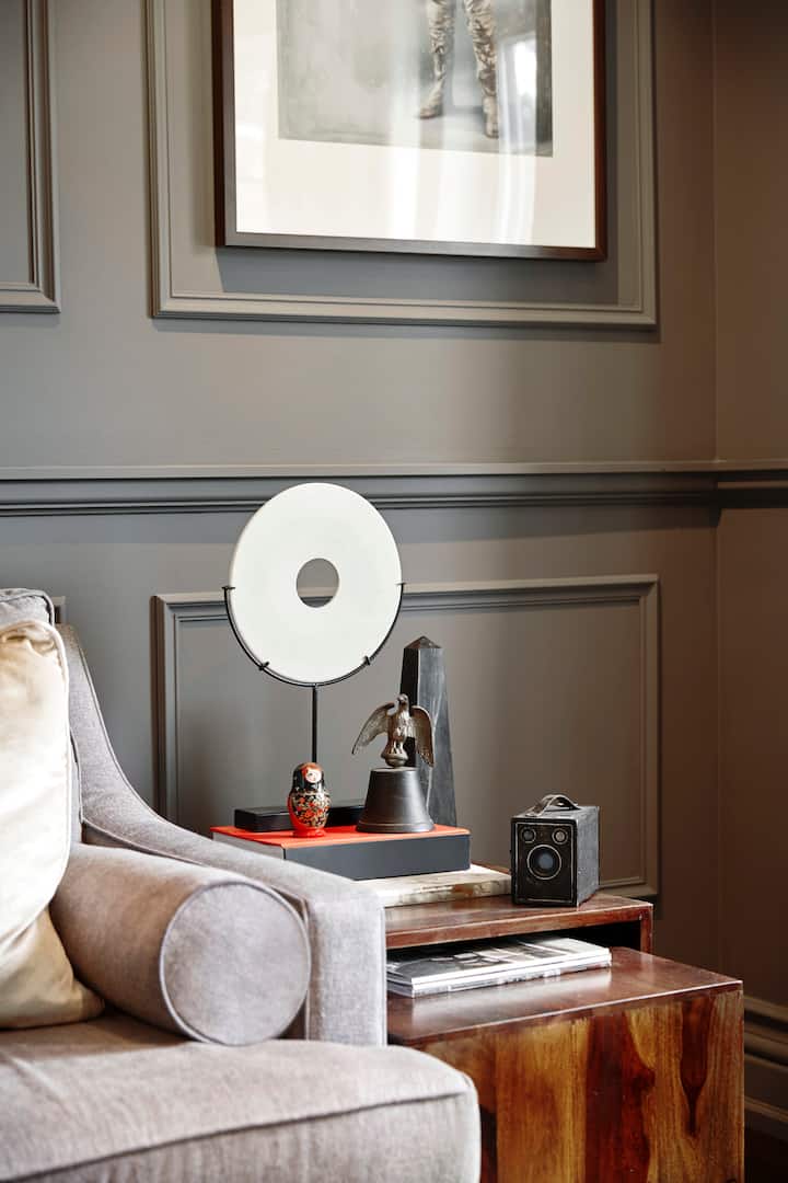 Dans le salon se trouve un mur à panneaux surmonté d'une œuvre d'art encadrée. Au premier plan, nous voyons le bord gauche d'un canapé gris, et une desserte en bois recouverte de livres, d'une caméra Brownie ancienne et de petits objets sculptés.