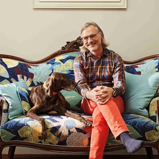 어깨 길이의 은발에 안경을 쓴 남성이 꽃무늬 소파에 갈색 개와 함께 앉아 미소를 짓고 있습니다.