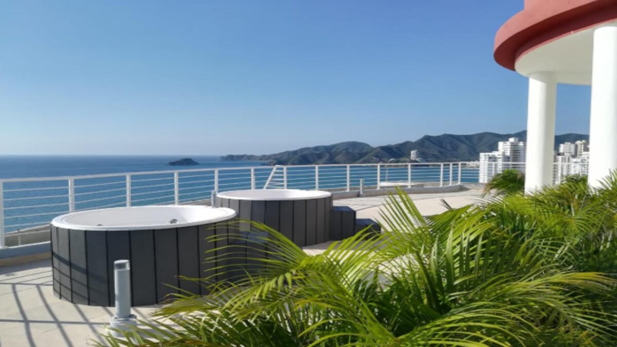 Two round jacuzzis are positioned on a spacious deck, surrounded by vibrant greenery. The Atlantic Ocean and distant mountains create a scenic backdrop under a clear blue sky, providing a tranquil atmosphere for relaxation.