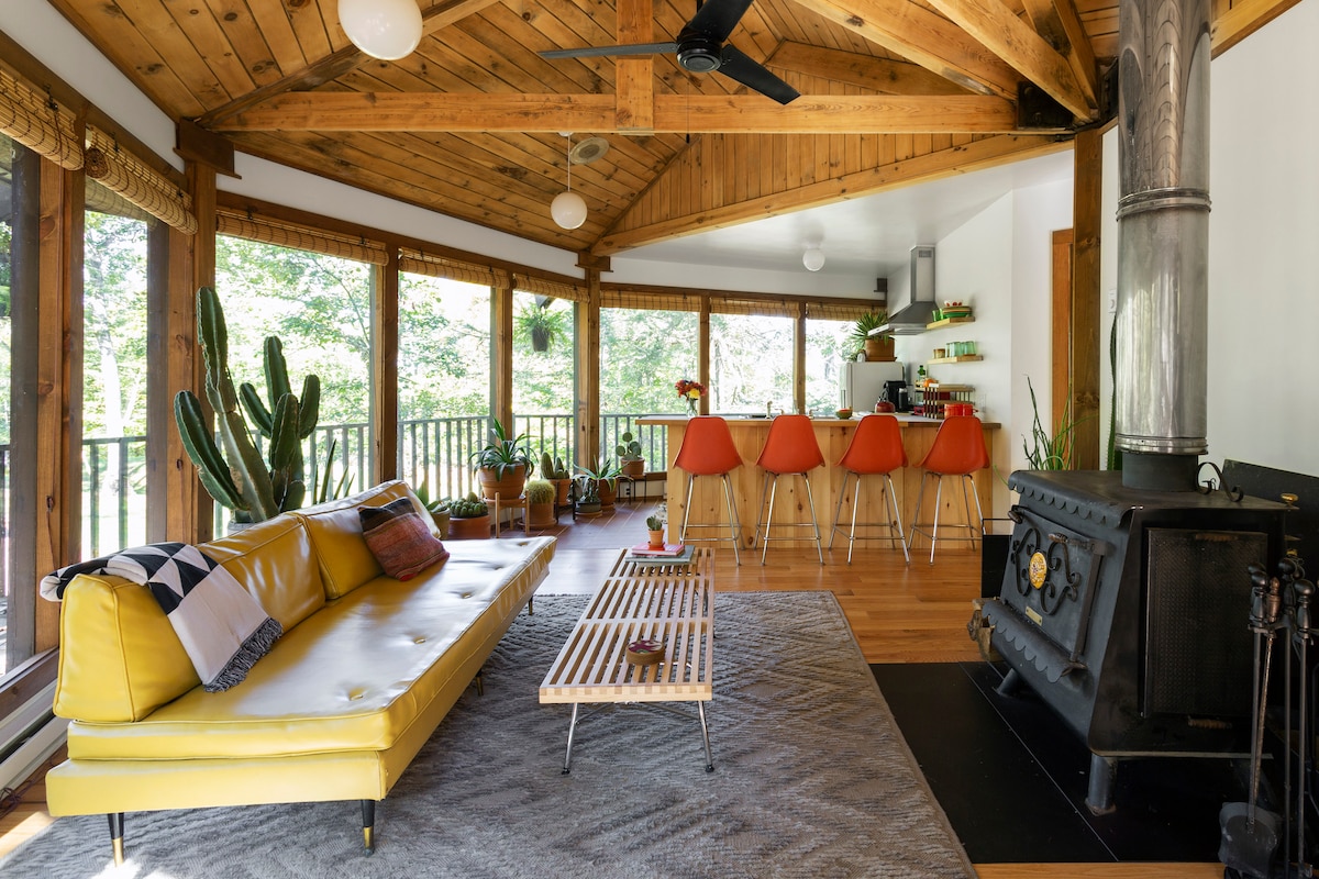 An open living area features a yellow sofa paired with a wooden coffee table. The space is enhanced by wood beams and large windows offering views of the surrounding greenery. A dining area with bright chairs is visible in the background, along with a well-equipped kitchen.