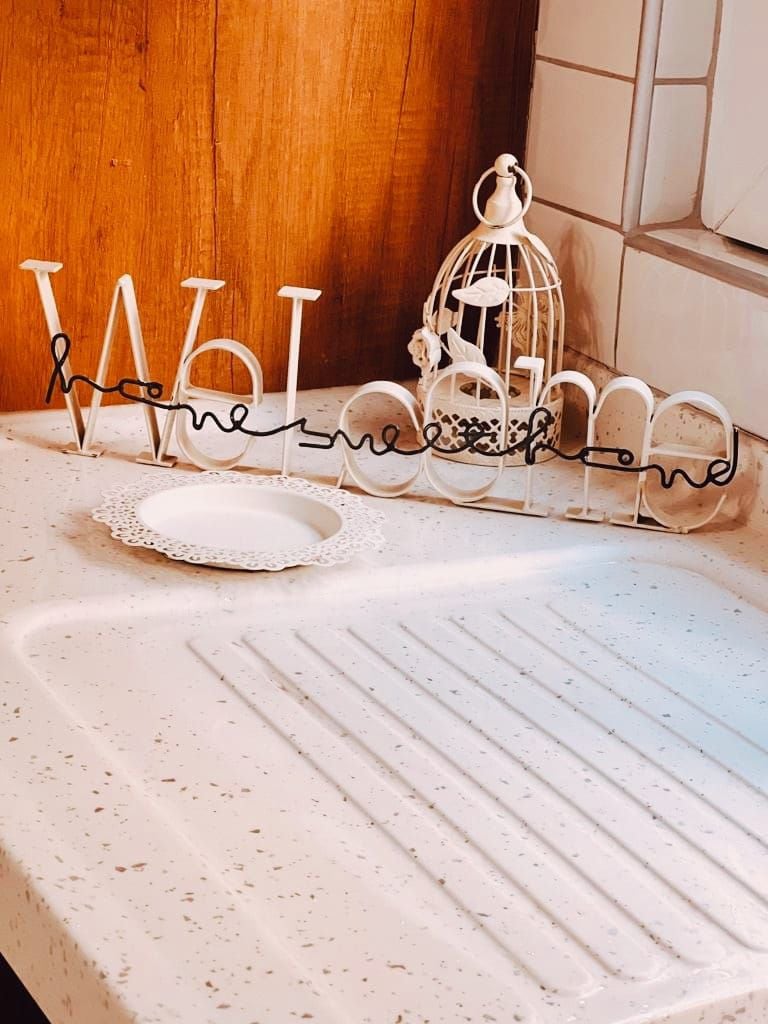 A kitchen countertop features a decorative sign that reads 'Welcome home' in white typography. A small, intricate plate rests nearby, complementing a vintage-style birdcage positioned in the background. Warm wood accents enhance the inviting atmosphere of the space.