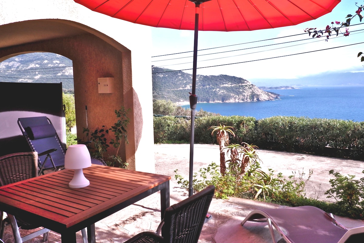 An outdoor terrace is depicted, featuring a wooden table with four chairs and a white lamp. A large red umbrella provides shade, while two lounge chairs are positioned nearby. The view of the sea and nearby hills is visible, creating a serene coastal ambiance.
