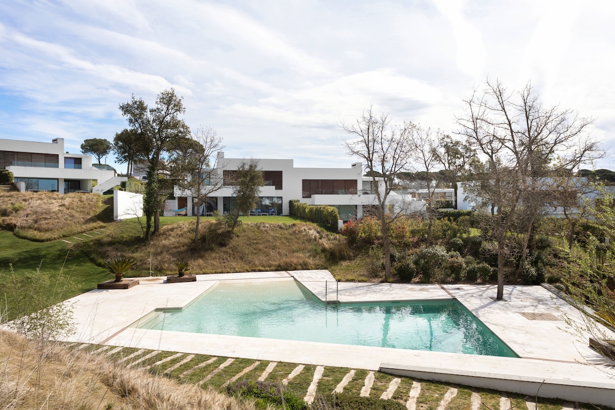 A serene outdoor space is depicted, featuring a private swimming pool surrounded by a manicured lawn. The pool area is complemented by gentle landscaping, with trees adding a natural element to the scene. The modern villa can be seen in the background, set against a bright sky.