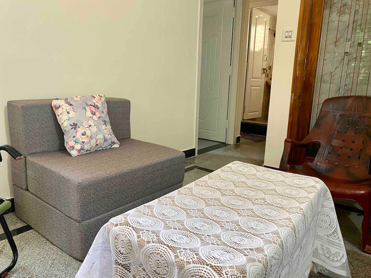 A cozy living area features a plush gray sofa bed adorned with a floral throw pillow. A delicate lace tablecloth covers a small coffee table, creating a homey atmosphere. The background reveals an entrance to the hallway with a visible door leading to additional spaces.