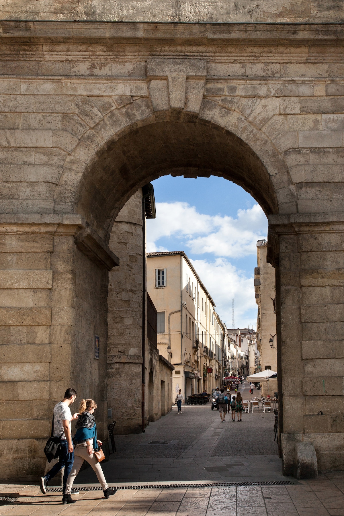 An ancient archway frames a lively street scene featuring people walking. The cobblestone path leads to buildings showcasing traditional architecture, while a blue sky and scattered clouds add a bright ambiance to the atmosphere.