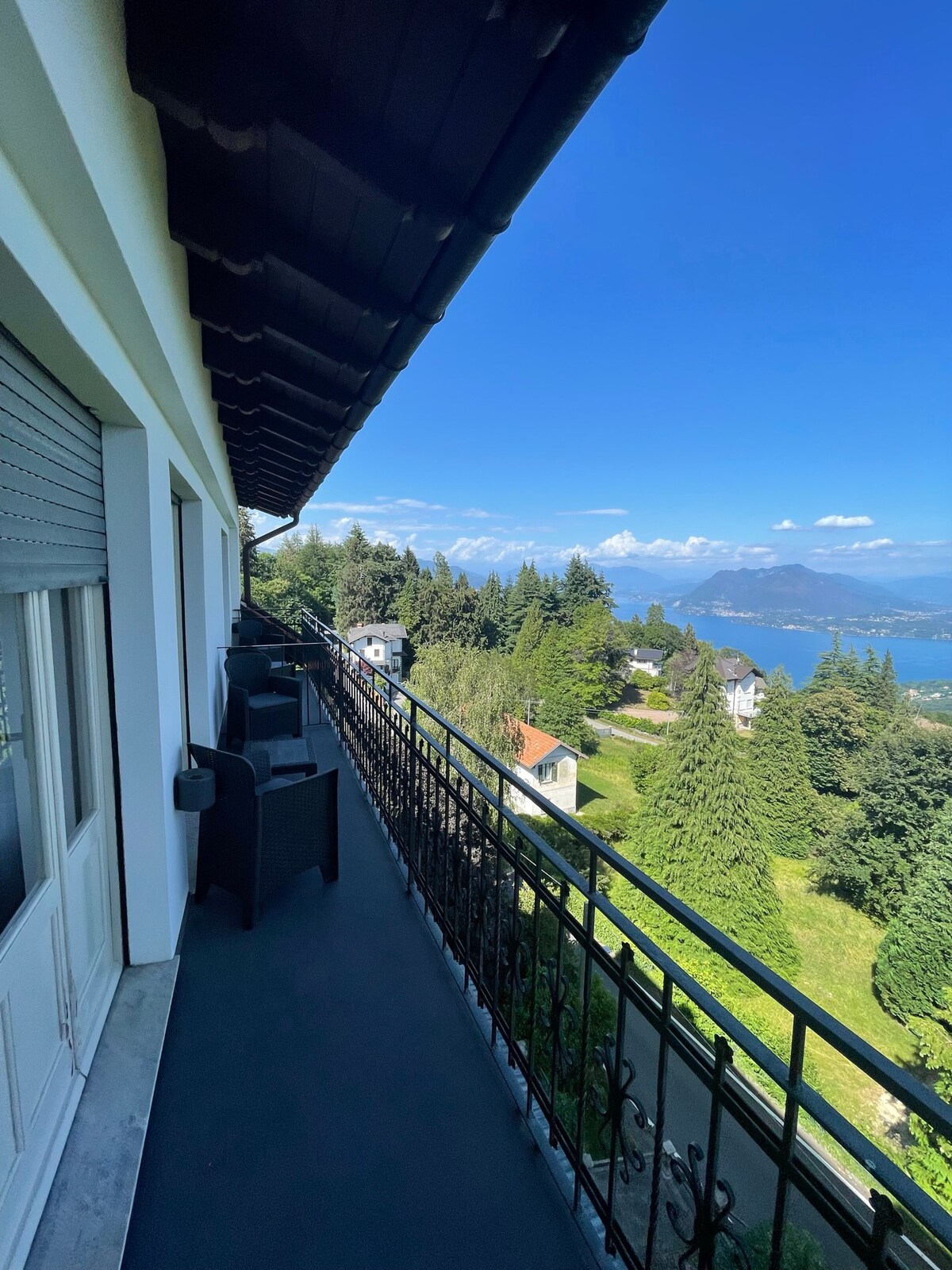 A balcony with a wrought iron railing offers views of lush greenery and distant hills. Two outdoor chairs are situated on the balcony, providing a relaxing spot to enjoy the surrounding landscape and the serene atmosphere.