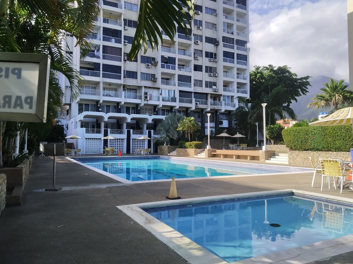 A large outdoor swimming pool area is visible, featuring two swimming pools surrounded by lounge chairs. The scene is framed by prominent tropical landscaping and a high-rise building in the background, with a bright sky enhancing the environment.