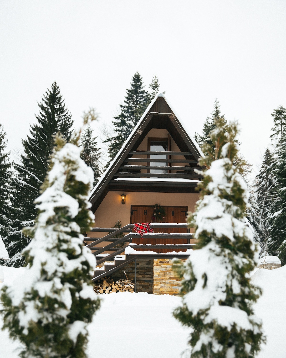 A charming mountain cottage is framed by snow-covered trees, showcasing a steeply pitched roof and wooden balconies. The front door, accented with decorative elements, is flanked by neatly stacked firewood beneath the porch, creating a welcoming entrance in a winter landscape.