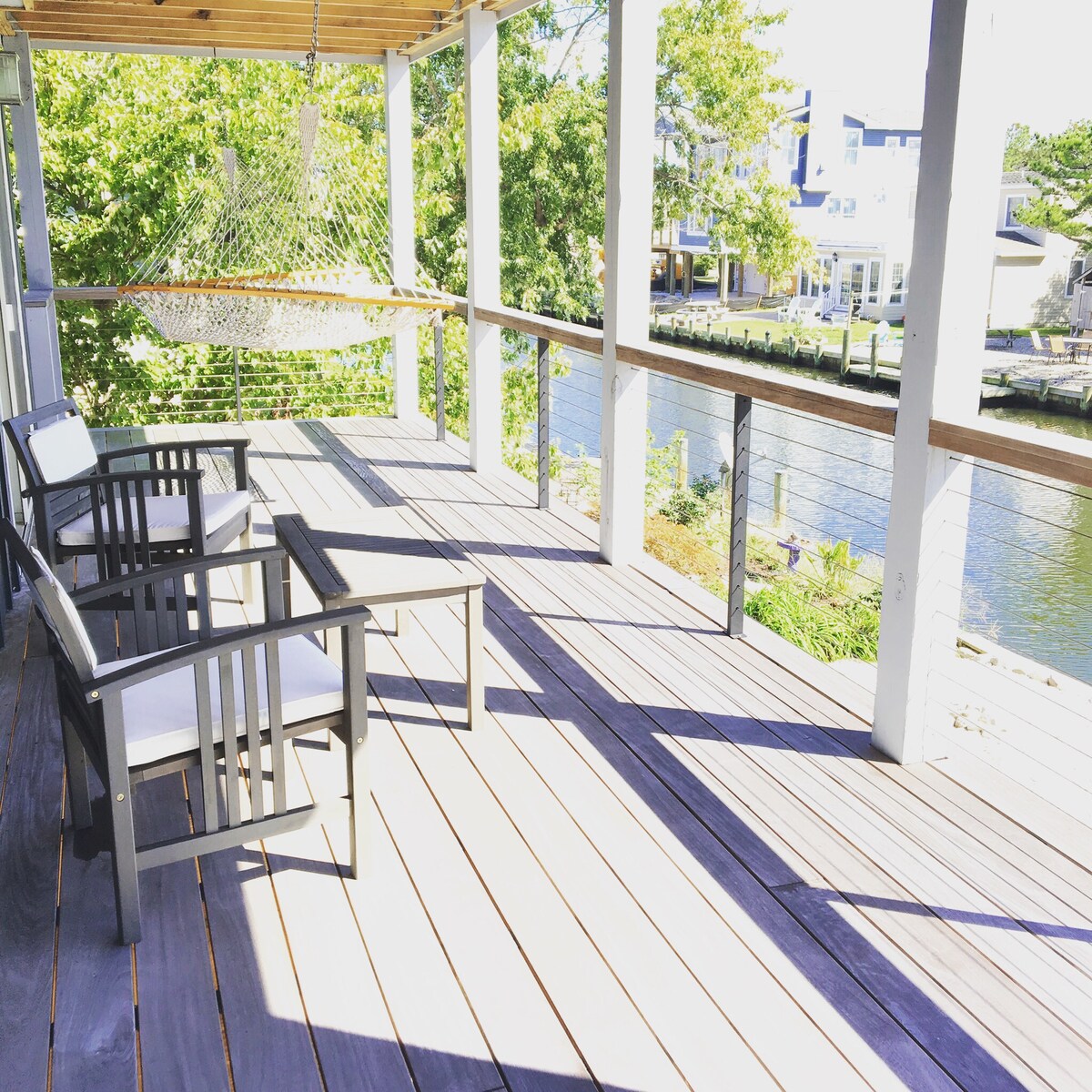 A shaded deck is shown featuring two modern outdoor chairs and a coffee table. A hammock hangs nearby, inviting relaxation. Sunlight casts soft shadows across the wooden flooring, while a tranquil canal is visible just beyond the railing, surrounded by greenery.