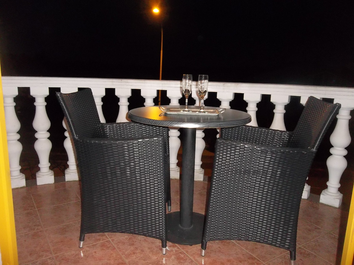 A small balcony table is paired with two woven chairs, positioned against a railing showcasing a classic balustrade. The setting features two glasses on the table, with a softly lit street lamp visible in the background, adding to the evening ambiance.