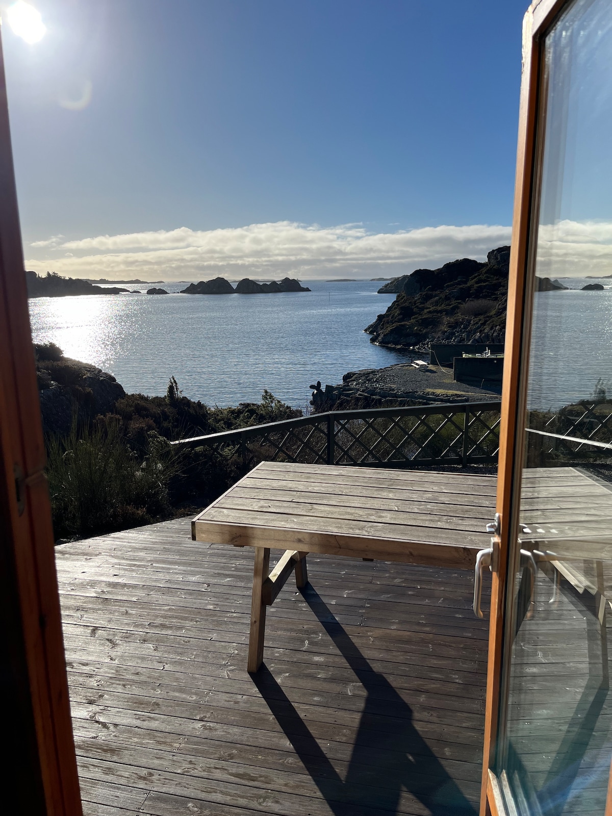 A spacious deck area is visible, featuring a wooden table positioned to face the calm waters. The scene captures a clear blue sky above, with distant rocky formations outlined by the sunlight reflecting off the water.