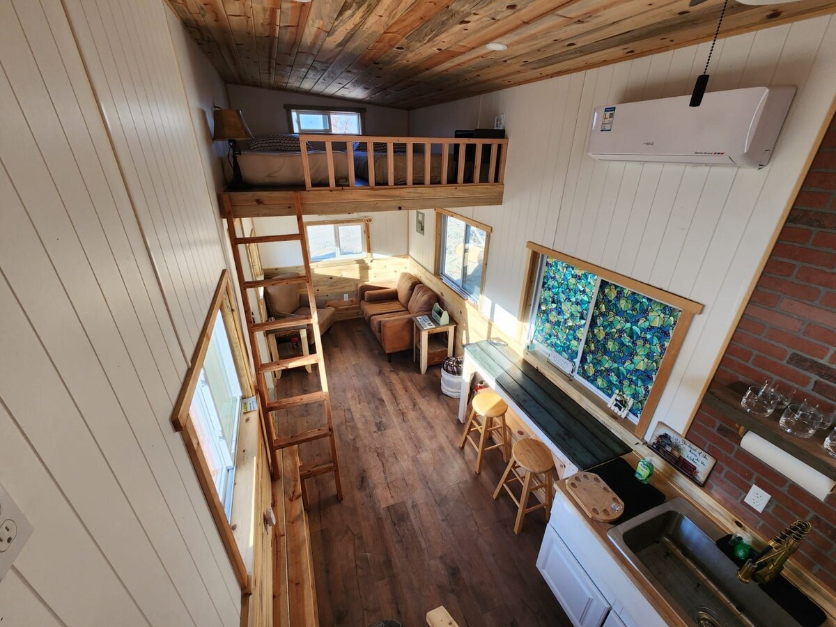 An aerial view of the tiny home's interior highlights a cozy layout. The master loft with a bed is visible above the sitting area, which includes a couch and two bar stools. Bright windows allow natural light to fill the space, showcasing wooden accents.