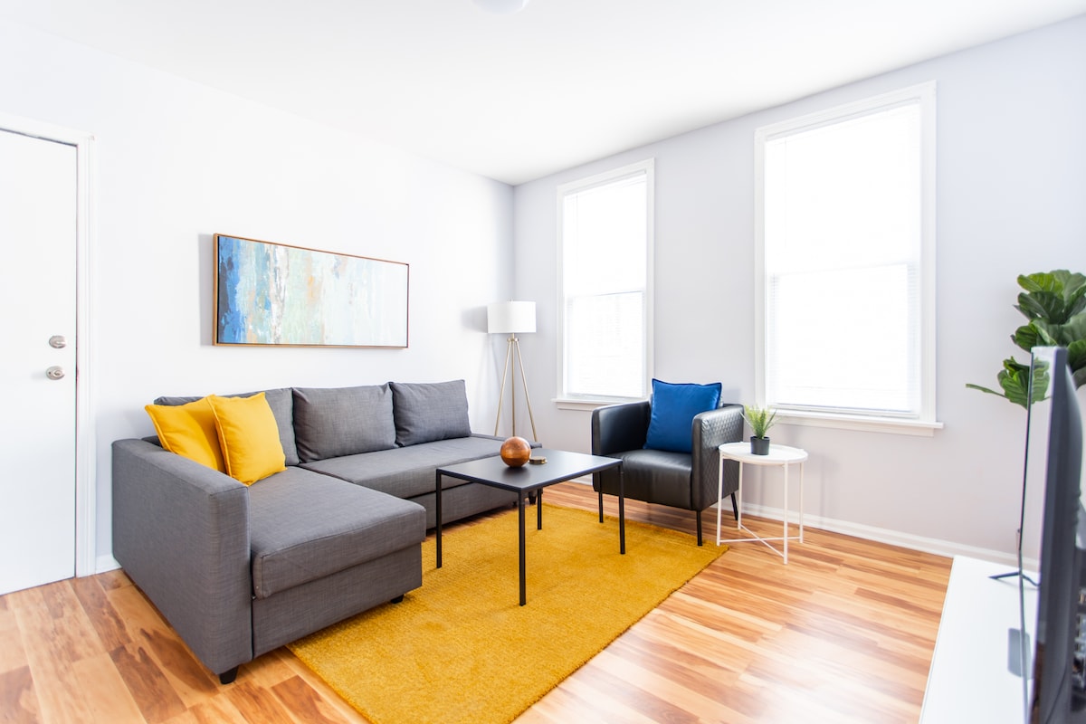 The living room features a plush sectional sofa adorned with bright yellow and blue accent pillows. A coffee table sits in front, complemented by a decorative bowl. Two windows allow ample natural light to stream in, illuminating the warm hardwood floors and contemporary art piece on the wall.