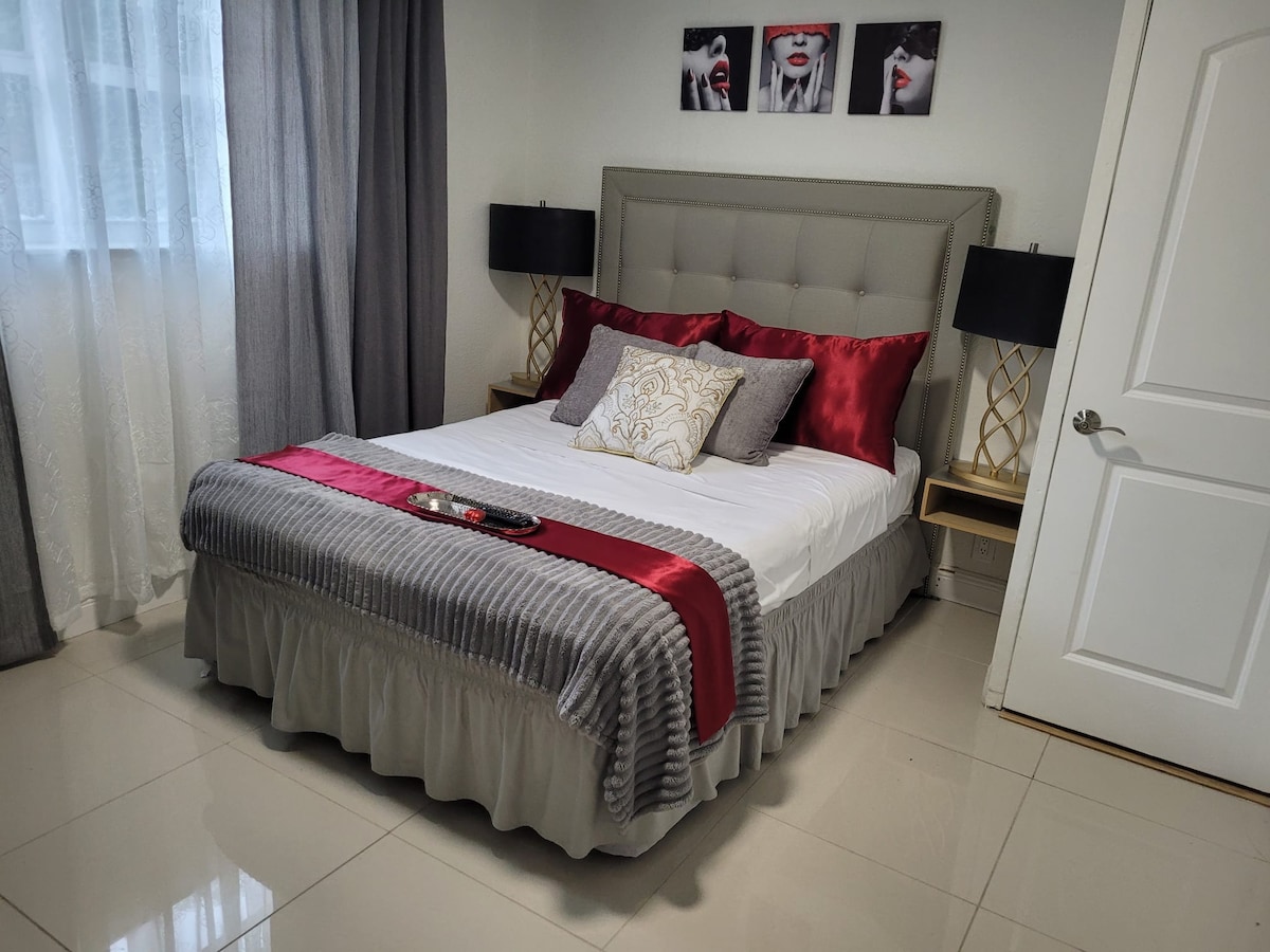 A cozy bedroom is furnished with a spacious bed dressed in soft linens. Elegant red and gray accents are featured in the bedding. Two bedside lamps complement the decor, while a series of framed artworks adorns the wall above the headboard. Natural light enters through a window with sheer curtains.