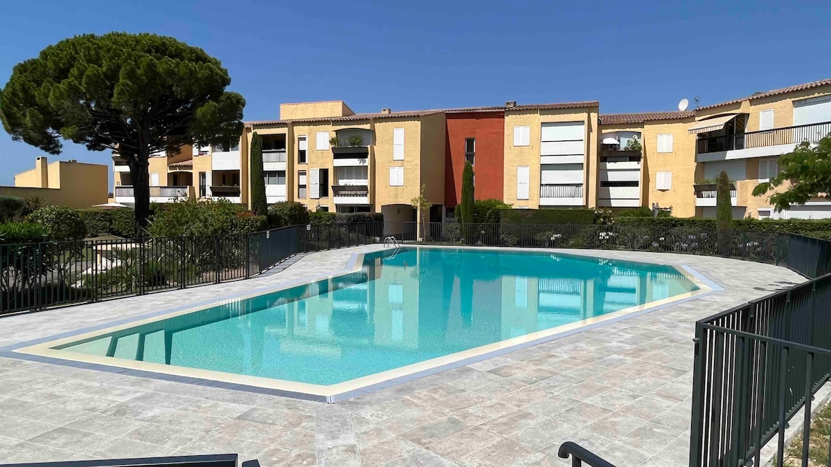 A large, inviting swimming pool is surrounded by a sun patio and a black railing. The water appears clear and well-maintained, reflecting the blue sky above. Colorful residential buildings are visible in the background, enhancing the peaceful outdoor setting.