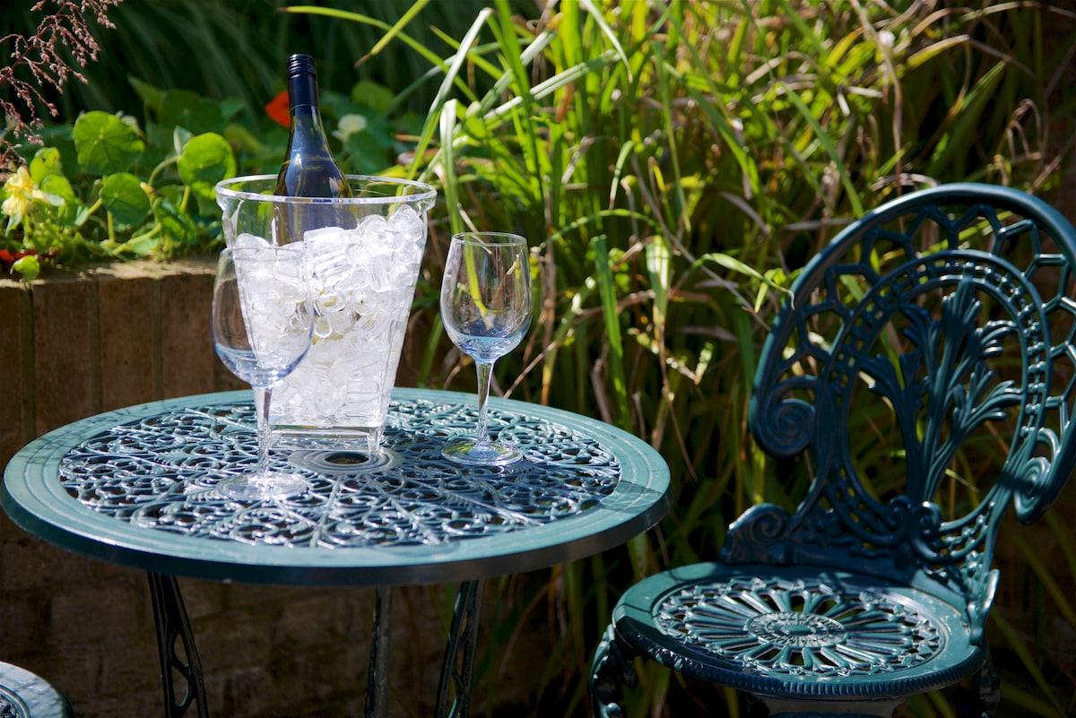 A small outdoor table is set with a wine bucket filled with ice, accompanied by two wine glasses. A decorative chair with intricate patterns complements the scene, surrounded by lush greenery, creating a serene garden atmosphere.