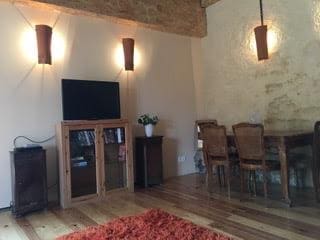 A spacious living area features a wooden floor and exposed stone walls. A flat-screen TV is mounted on the wall above a cabinet, while a dining table with wicker chairs is positioned alongside. Soft lighting from wall sconces creates a relaxed ambiance.