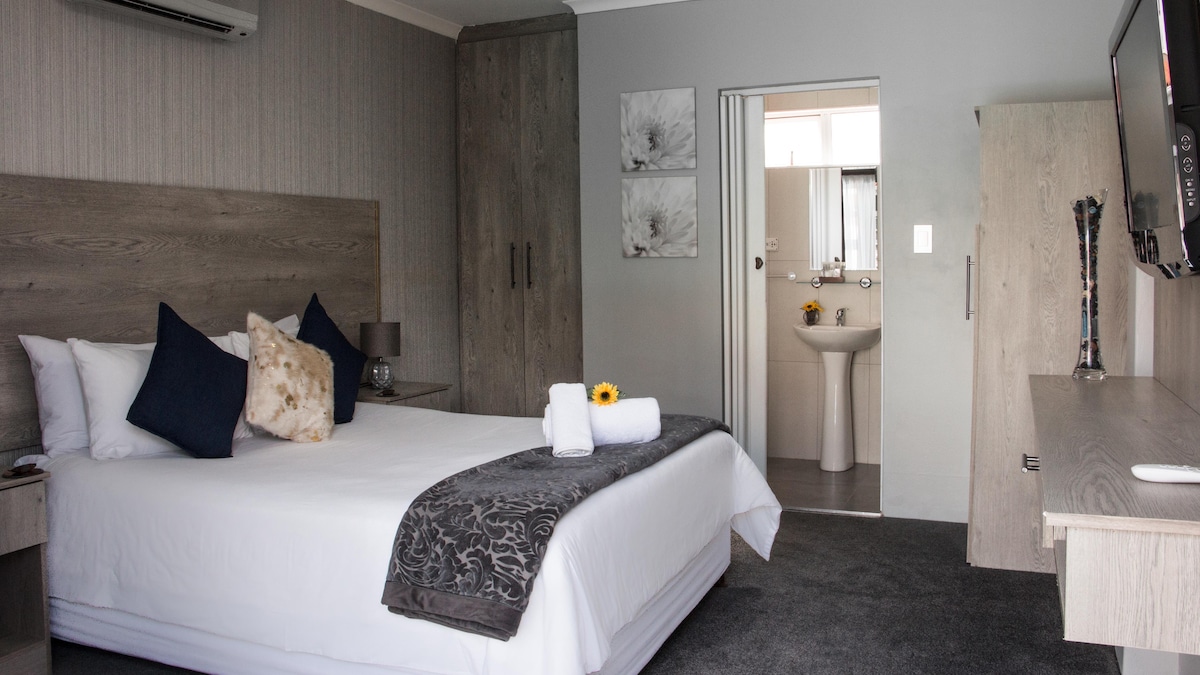 A well-appointed guest room features a spacious bed adorned with white linens and accent pillows. Natural light enters through the adjacent bathroom door, illuminating the room. A flat-screen television and a vanity area are positioned within easy reach, contributing to a comfortable environment.