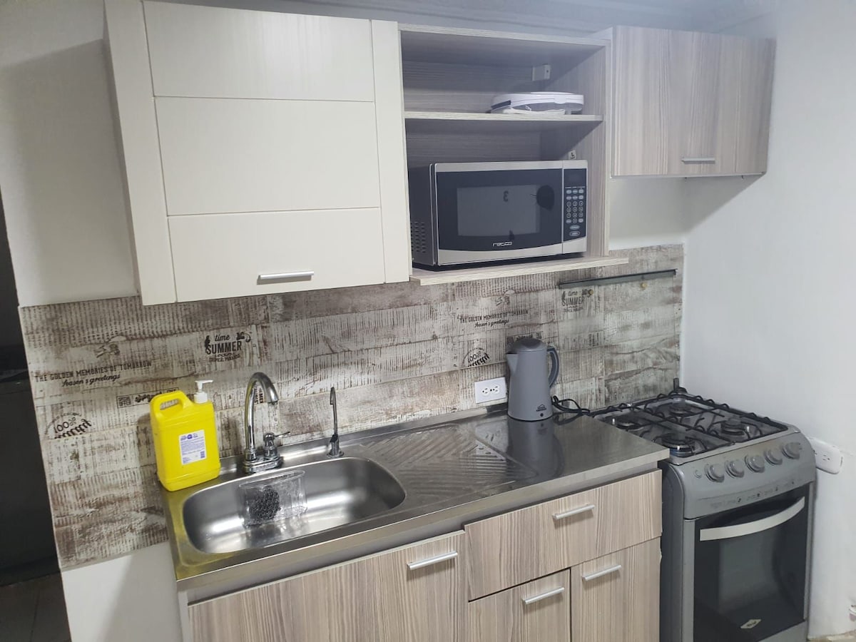 The kitchen features modern appliances, including a microwave and gas stove. A stainless steel sink is positioned beneath upper cabinetry, which includes storage space. A stylish backsplash adds texture to the overall design, complementing the light wood cabinetry.