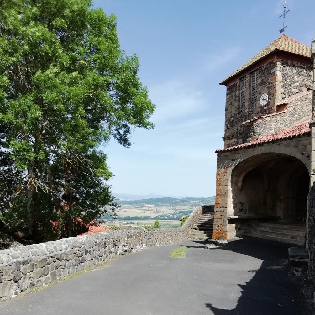 A stone building with a clock tower is visible, framed by a large green tree. The pathway leads to steps that descend beside the structure, offering a view of the hills and fields in the background under a clear blue sky.