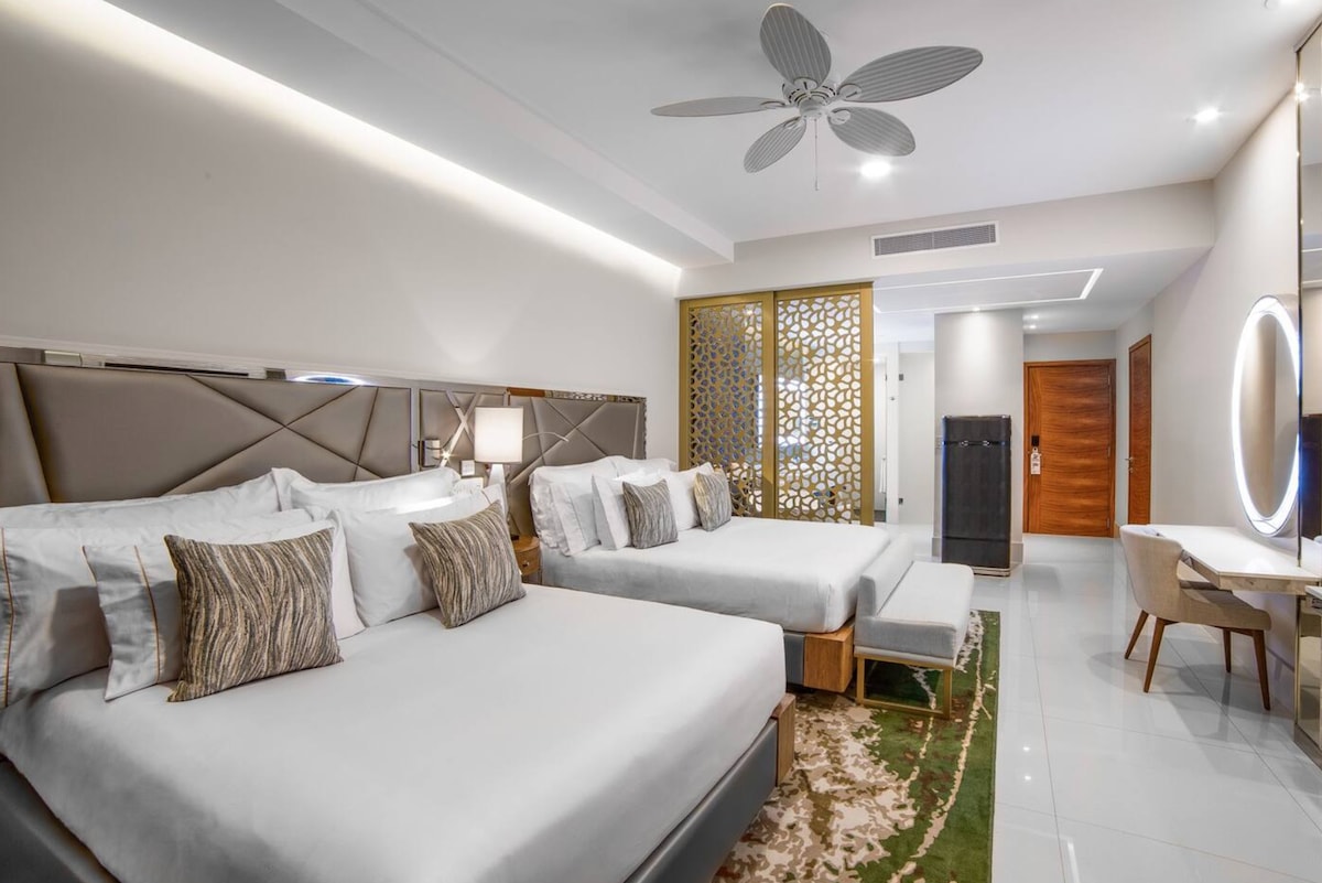 A spacious suite features two queen beds adorned with decorative pillows. A neutral color palette is complemented by a stylish screen and a modern desk area. Soft lighting and a ceiling fan enhance the room's ambiance, while sleek flooring contributes to a polished look.