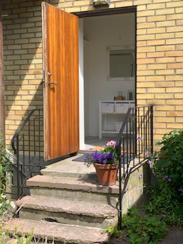 A wooden front door stands open, revealing a light-filled entrance. Stone steps lead up to the door, framed by a small iron railing. A potted plant with colorful flowers sits on the step, adding a touch of greenery to the welcoming entry.