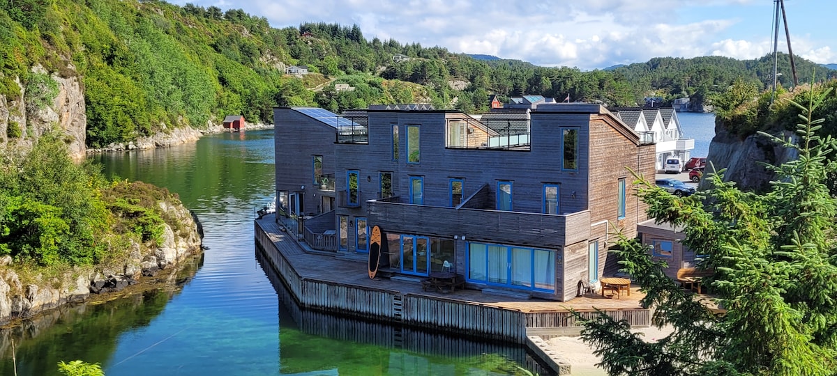 The modern building is situated on the water's edge, surrounded by lush greenery. Large windows and sliding glass doors provide scenic views of the serene surroundings. A wooden deck extends from the structure, offering outdoor seating and direct access to the water.
