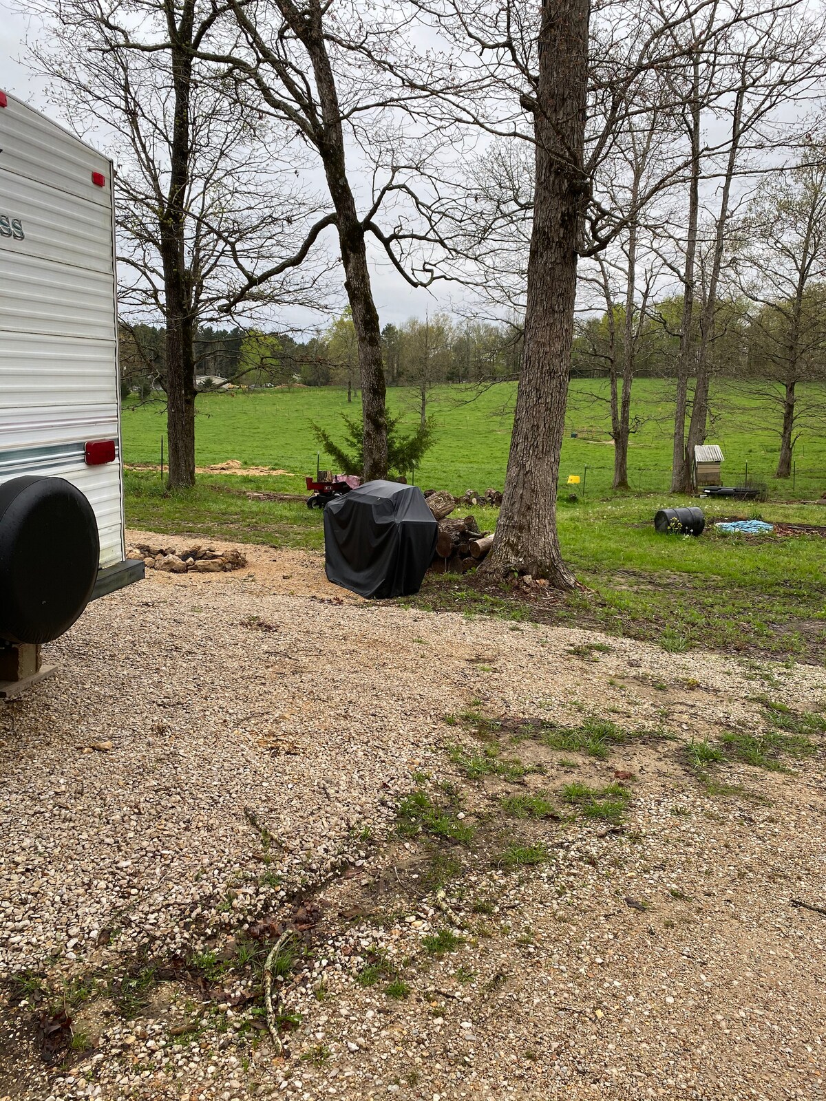 A gravel area is shown next to a camper. A black grill cover is visible near a tree, surrounded by a grassy lawn with scattered colorful objects in the distance. The landscape features several trees and open green space.