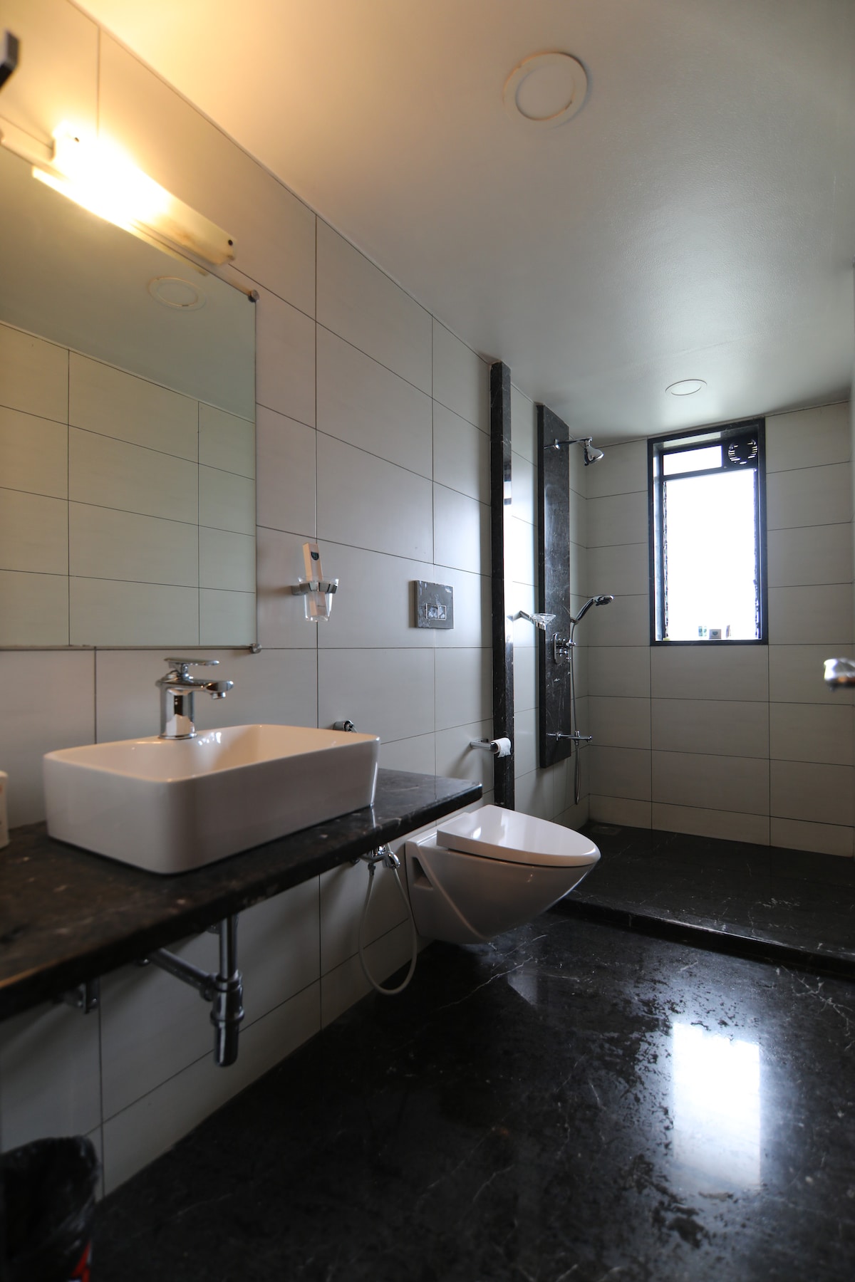 The modern bathroom features a sleek design with light-colored tiles and a spacious shower area. A large mirror is mounted above a minimalist sink, complemented by a dark granite countertop. Natural light enters through a tall window, enhancing the contemporary feel.