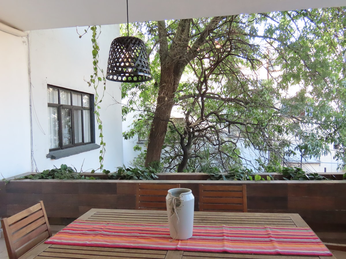 A wooden dining table is positioned on a covered terrace, accompanied by matching chairs. A colorful striped runner is placed on the table, while a simple ceramic container sits in the center. A large tree is visible beyond the railing, surrounded by greenery.