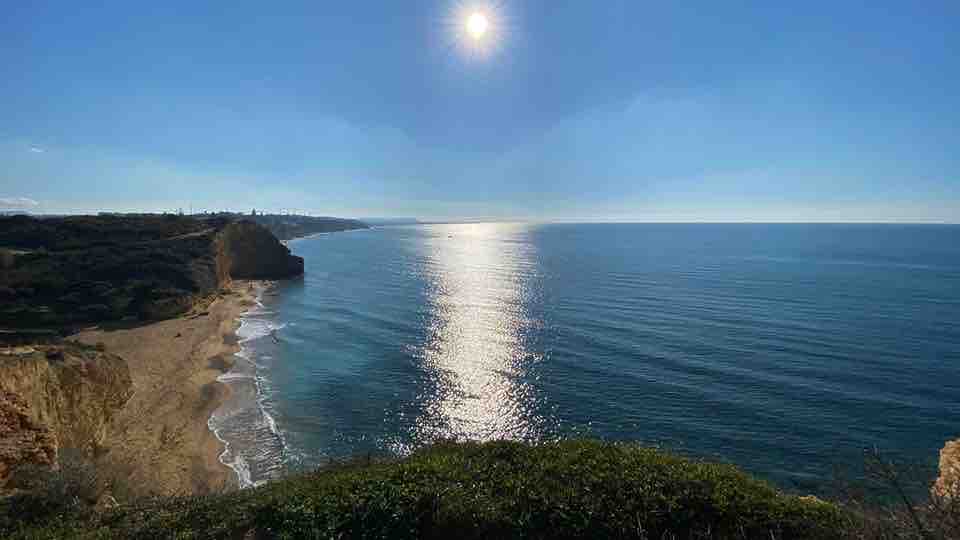 A coastal view is captured, featuring a sunlit sea that reflects the bright sky. The shoreline is visible in the distance, framed by gentle cliffs and greenery, creating a serene landscape. The image conveys a sense of tranquility and natural beauty.