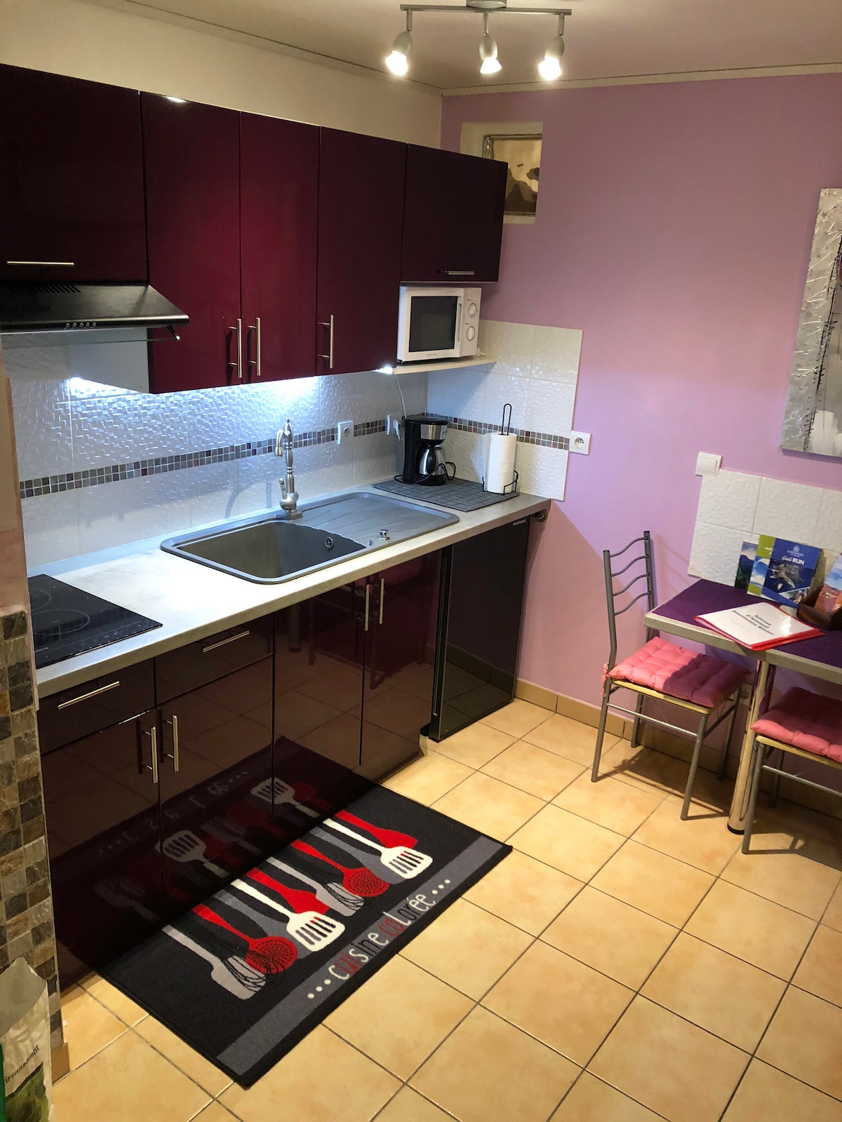 The kitchen features modern dark cabinetry and a stylish backsplash. A microwave, coffee maker, and kitchen utensils are provided. A small dining area is present with a table and two chairs, complemented by a vibrant area rug showcasing utensils. Soft lighting enhances the space.