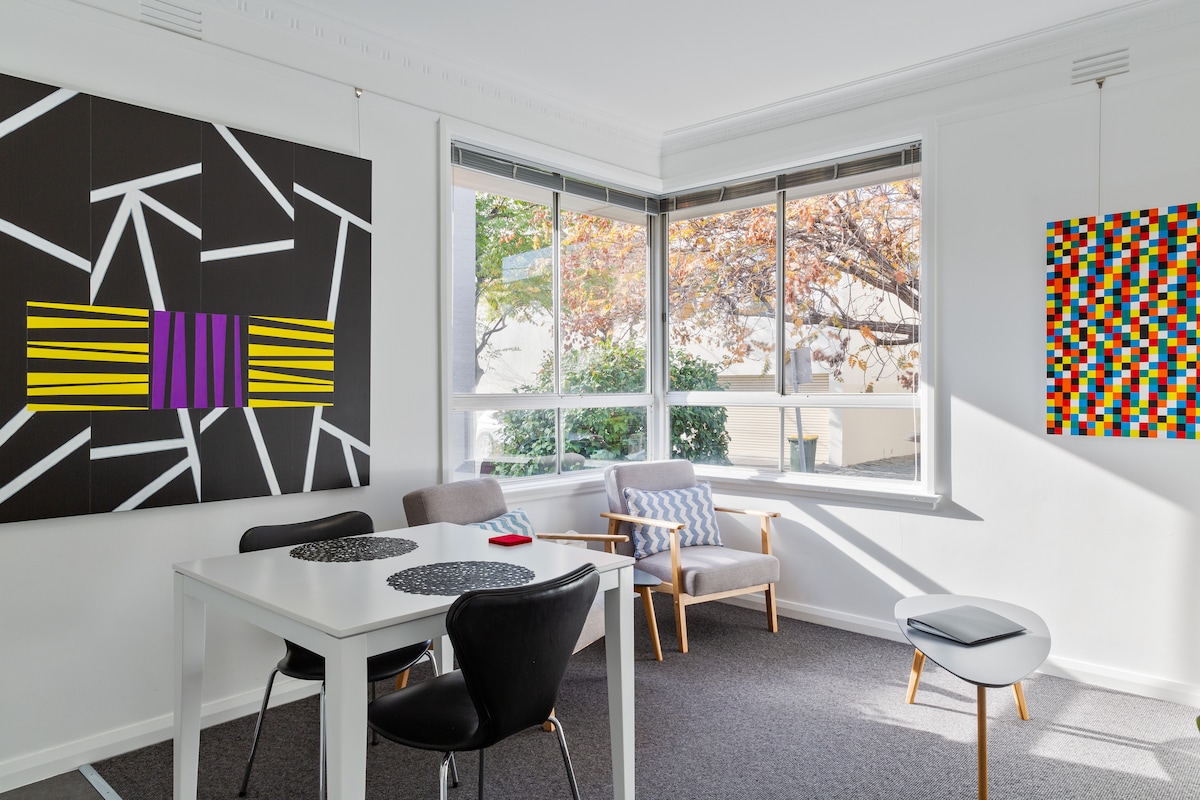 A bright living area features large windows allowing natural light, with views of leafy surroundings. A dining table is set for four, complemented by a comfortable chair. Two colorful artworks adorn the walls, adding a modern touch to the space.
