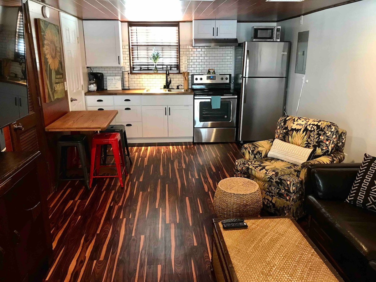 The interior living space features an open kitchen with stainless steel appliances and a breakfast bar with three high stools. A cozy seating area includes a floral-patterned armchair and a dark leather couch. Natural light enters through the window, highlighting the wood-style flooring.