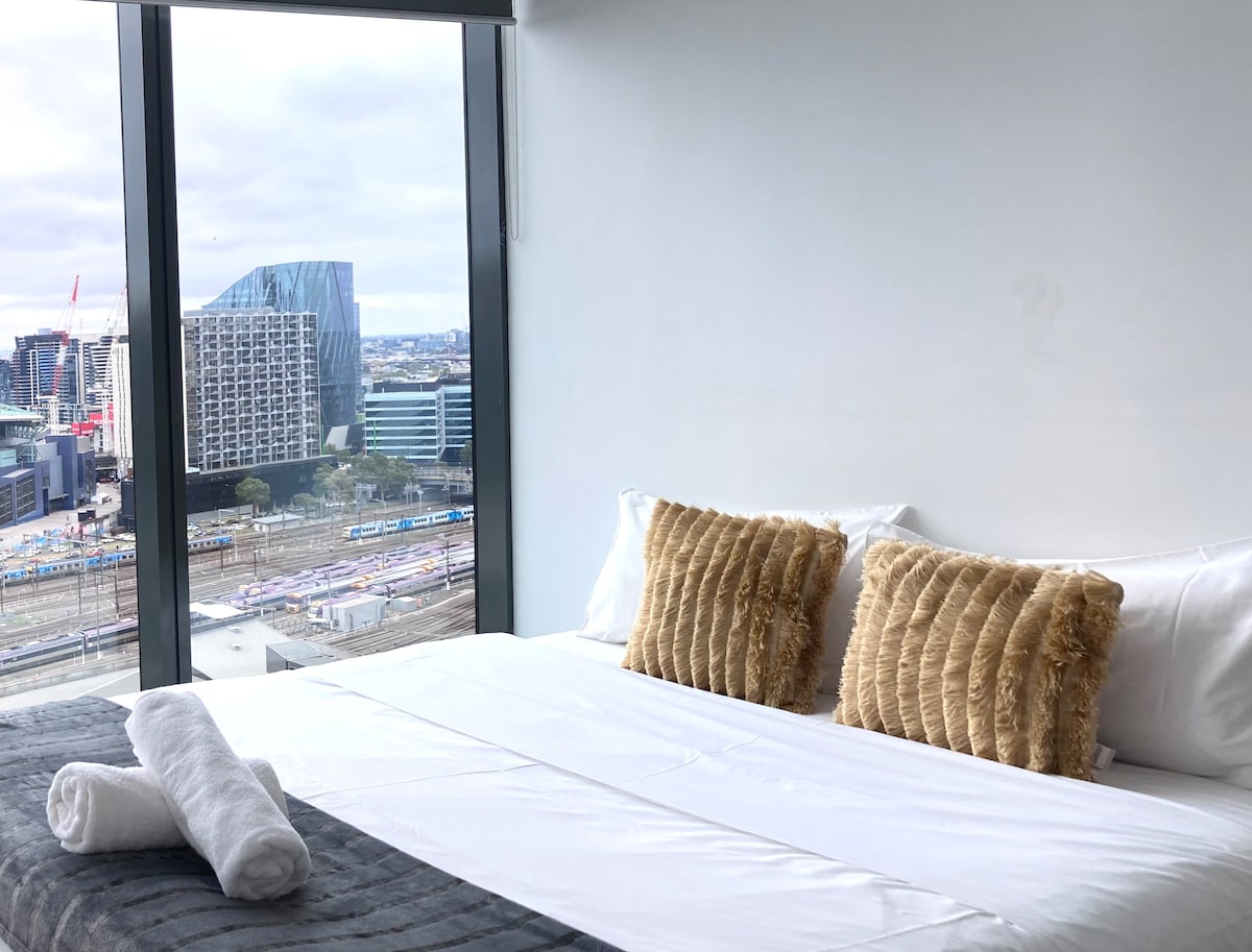A spacious bedroom features a large bed adorned with plush, textured pillows and fresh linens. Floor-to-ceiling windows provide a panoramic view of the city skyline and train tracks below, allowing natural light to fill the room.