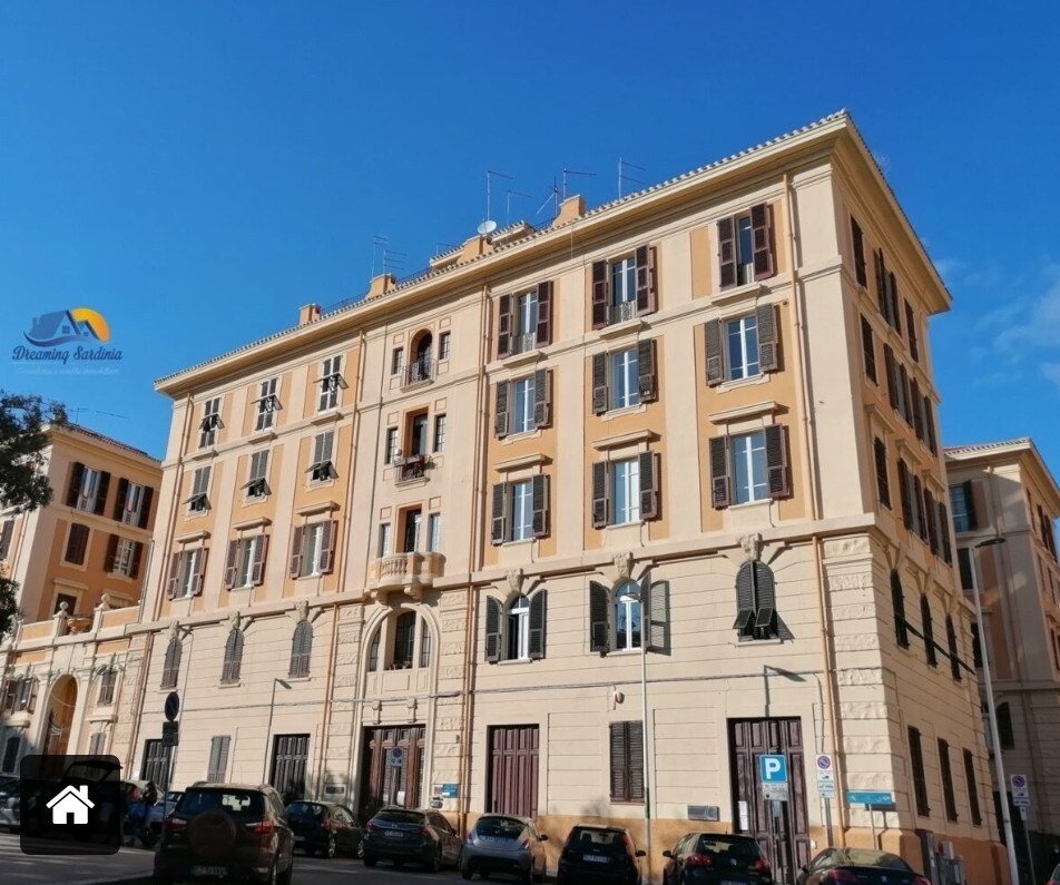 The exterior of the elegant palazzo features a classic architectural design, characterized by a light-colored façade and multiple windows. A well-maintained entrance is visible at the ground level, surrounded by a vibrant urban environment with nearby vehicles and clear blue skies above.
