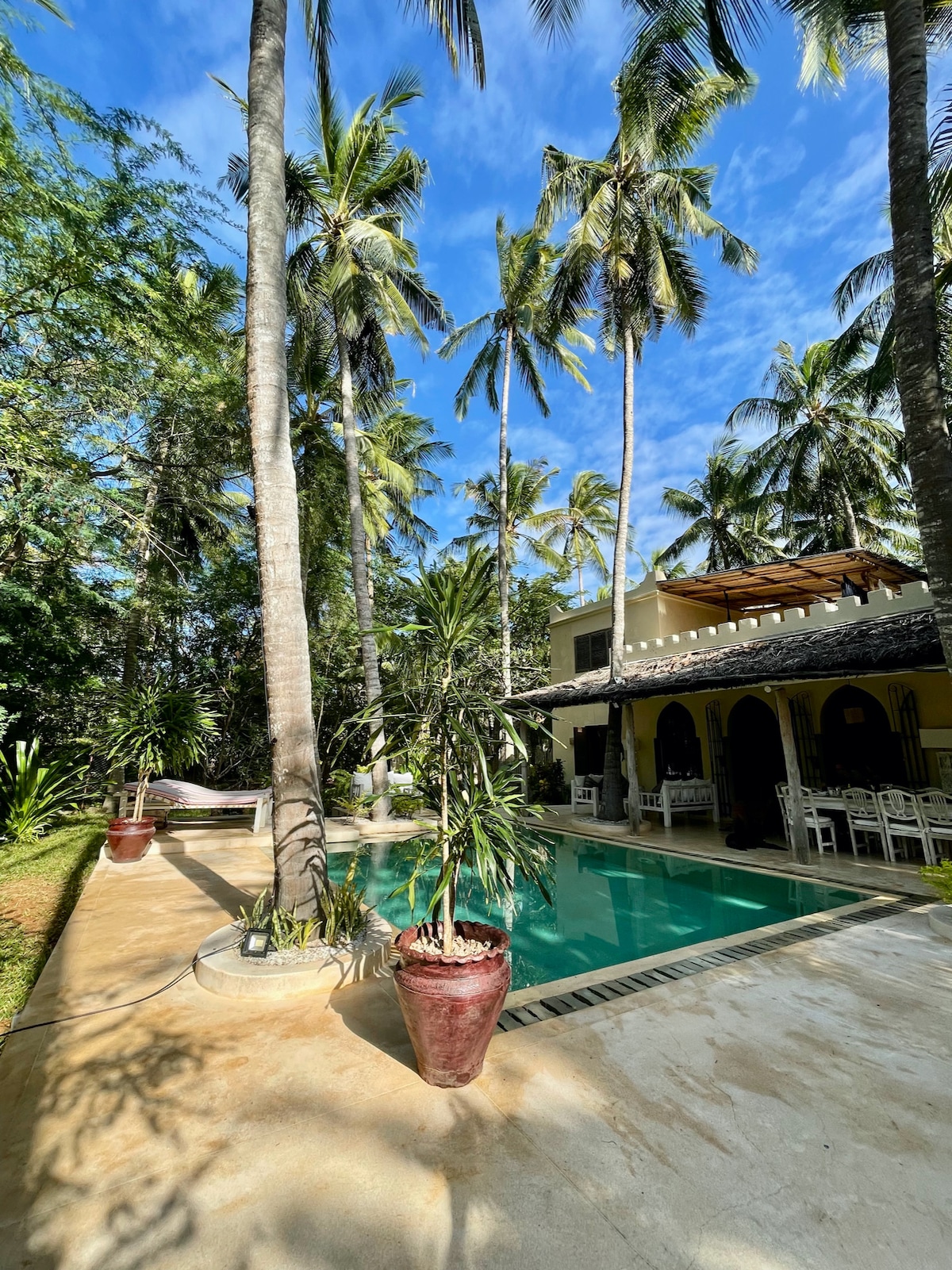A private pool is surrounded by tall palm trees and lush greenery. The pool area features a smooth stone deck and potted plants, while the house's open structure invites natural light and views of the outdoor space. A cozy seating area is visible nearby.