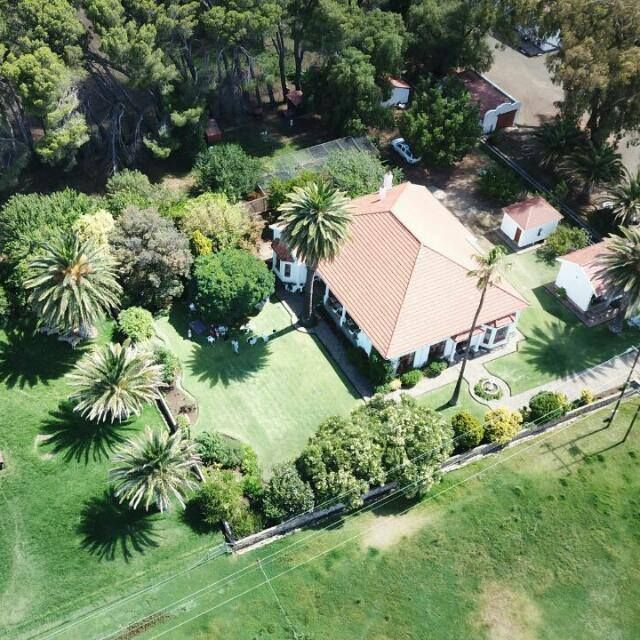 An aerial view of the property showcases a spacious home surrounded by lush greenery and palm trees. The well-maintained lawn is dotted with various trees and flower beds, creating a serene atmosphere. Nearby structures and open fields are also visible, suggesting a rural setting.