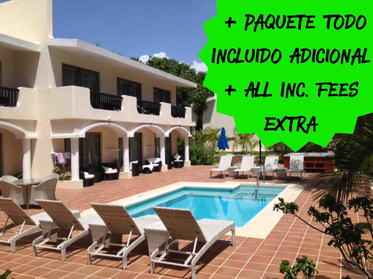 A tranquil courtyard features a central swimming pool surrounded by elegant loungers and chairs. Archways connect to rooms with balconies overlooking the pool area. Lush greenery provides a serene backdrop, enhancing the relaxing atmosphere of the space.