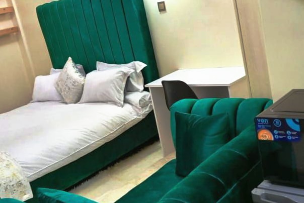 A cozy sleeping area is furnished with a spacious bed featuring a plush green headboard. A small desk and chair are positioned nearby, complemented by a soft couch in the same rich green tone, creating a harmonious and inviting environment.
