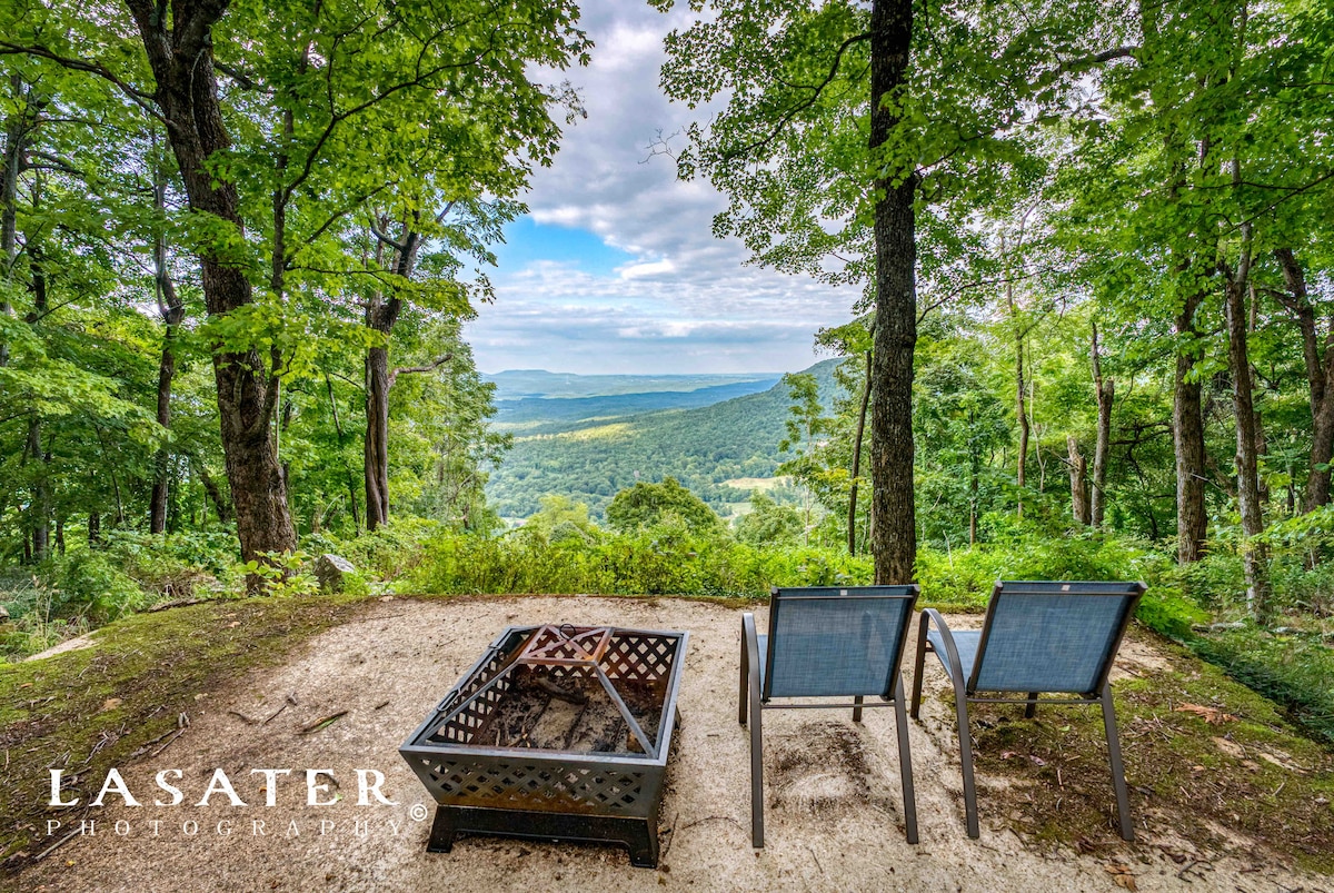 Two metal chairs are positioned around a fire pit on a gravel surface amidst a lush forest. An expansive view of rolling hills and trees is visible in the background, with a cloudy sky overhead, creating a serene outdoor setting.