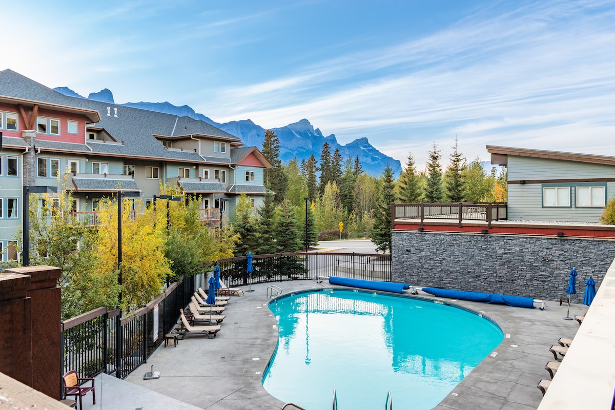 A spacious outdoor pool is surrounded by lounge chairs, with a backdrop of trees and distant mountains. The clear blue water reflects the sky, creating a serene atmosphere. The well-maintained area is enclosed by fencing, ensuring a comfortable environment for guests.